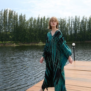 May include: A person wearing a long, flowing, black and teal tie-dye dress, standing on a wooden dock over a lake. The person is looking at the camera.