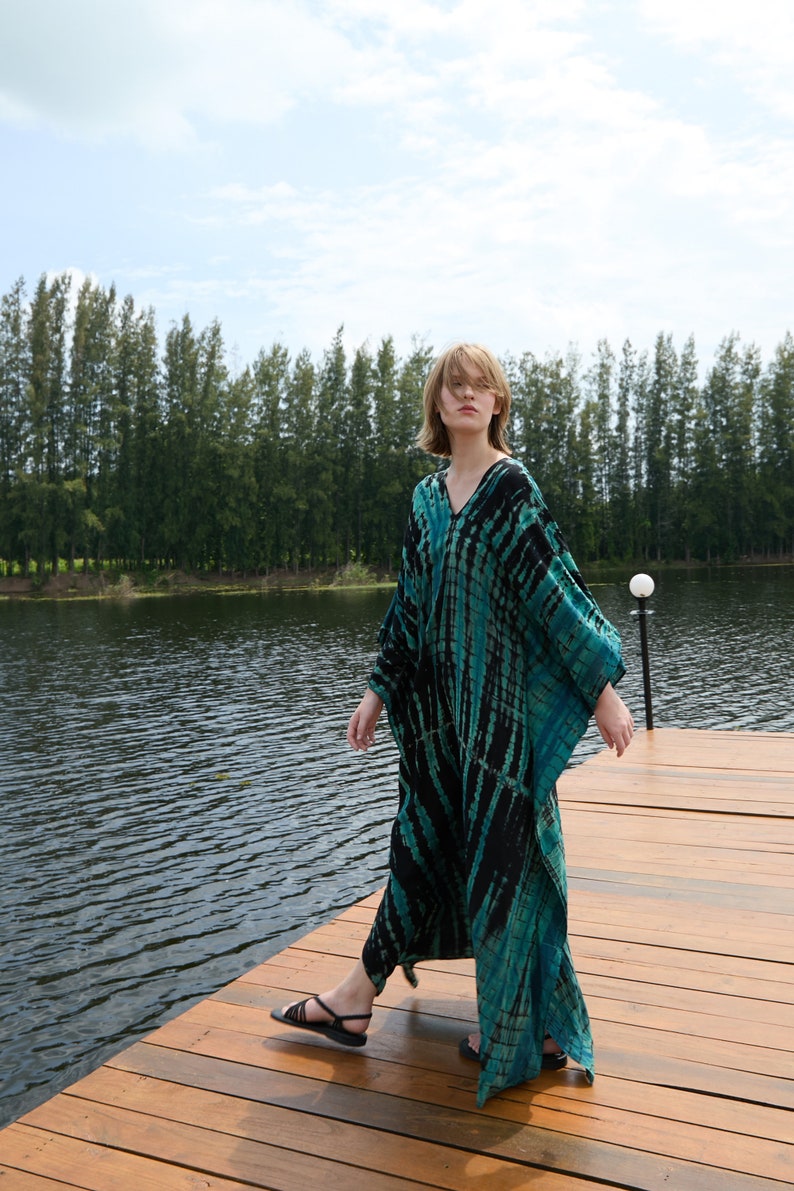 May include: A person wearing a long, flowing, black and teal tie-dye dress, standing on a wooden dock over a lake. The person is looking at the camera.