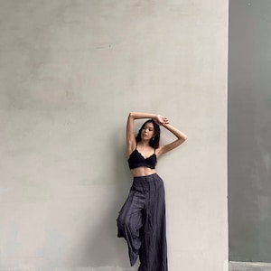 Puede incluir: Una mujer con un sujetador de encaje negro y pantalones anchos de color gris oscuro se encuentra de pie frente a una pared blanca.