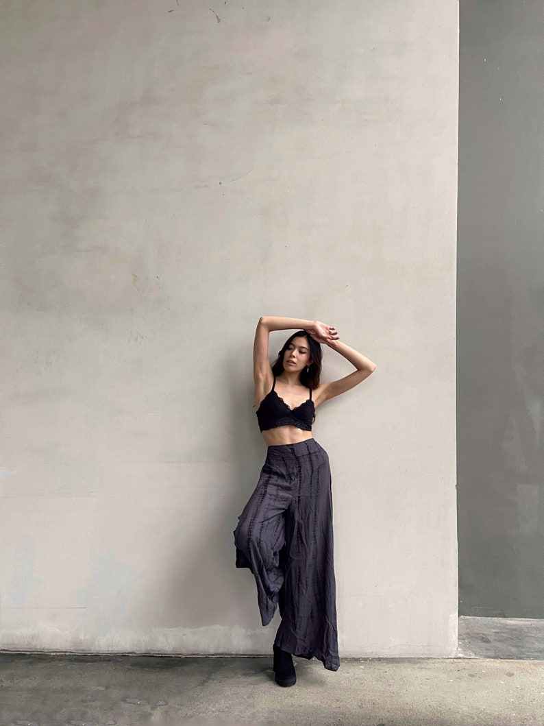 Puede incluir: Una mujer con un sujetador de encaje negro y pantalones anchos de color gris oscuro se encuentra de pie frente a una pared blanca.