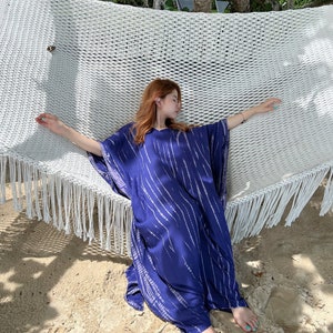 May include: A woman's blue caftan with white vertical stripes is draped over a white hammock. The caftan has a V-neck and wide sleeves. The hammock is suspended between trees on a sandy beach.