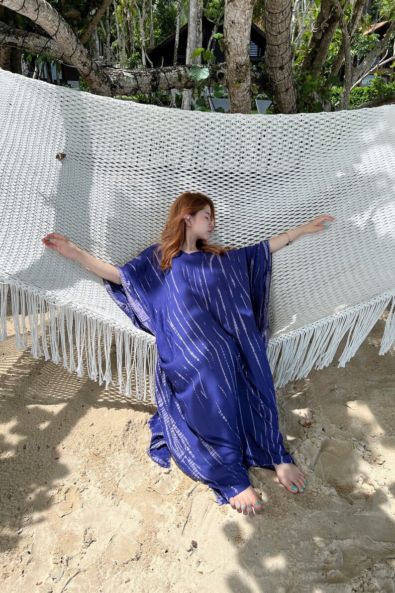 May include: A woman's blue caftan with white vertical stripes is draped over a white hammock. The caftan has a V-neck and wide sleeves. The hammock is suspended between trees on a sandy beach.