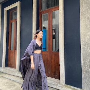 Puede incluir: Una mujer con un sujetador negro y un traje de pantal&oacute;n largo y fluido, de color morado y gris tie-dye. Est&aacute; caminando por un callej&oacute;n estrecho con puertas de madera a ambos lados.