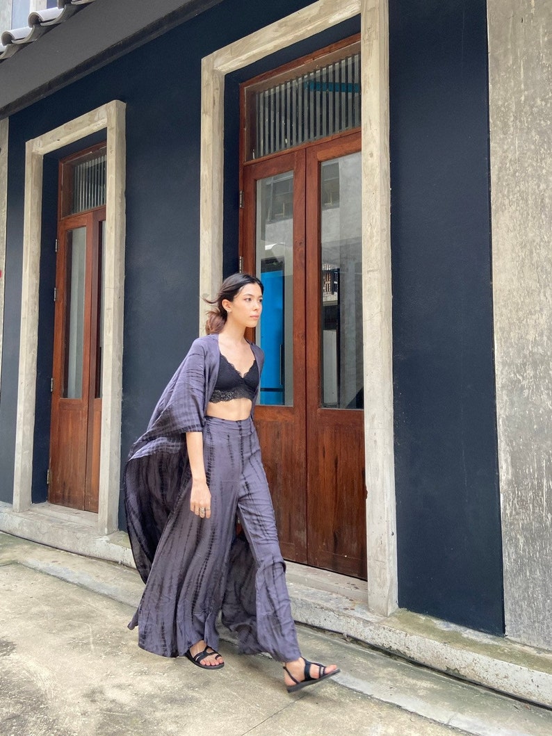 Puede incluir: Una mujer con un sujetador negro y un traje de pantal&oacute;n largo y fluido, de color morado y gris tie-dye. Est&aacute; caminando por un callej&oacute;n estrecho con puertas de madera a ambos lados.