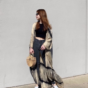 May include: A woman wearing a long, flowing tie-dye kimono with black accents over a black crop top and dark wash jeans. She is carrying a small, round, woven bag and wearing white shoes.