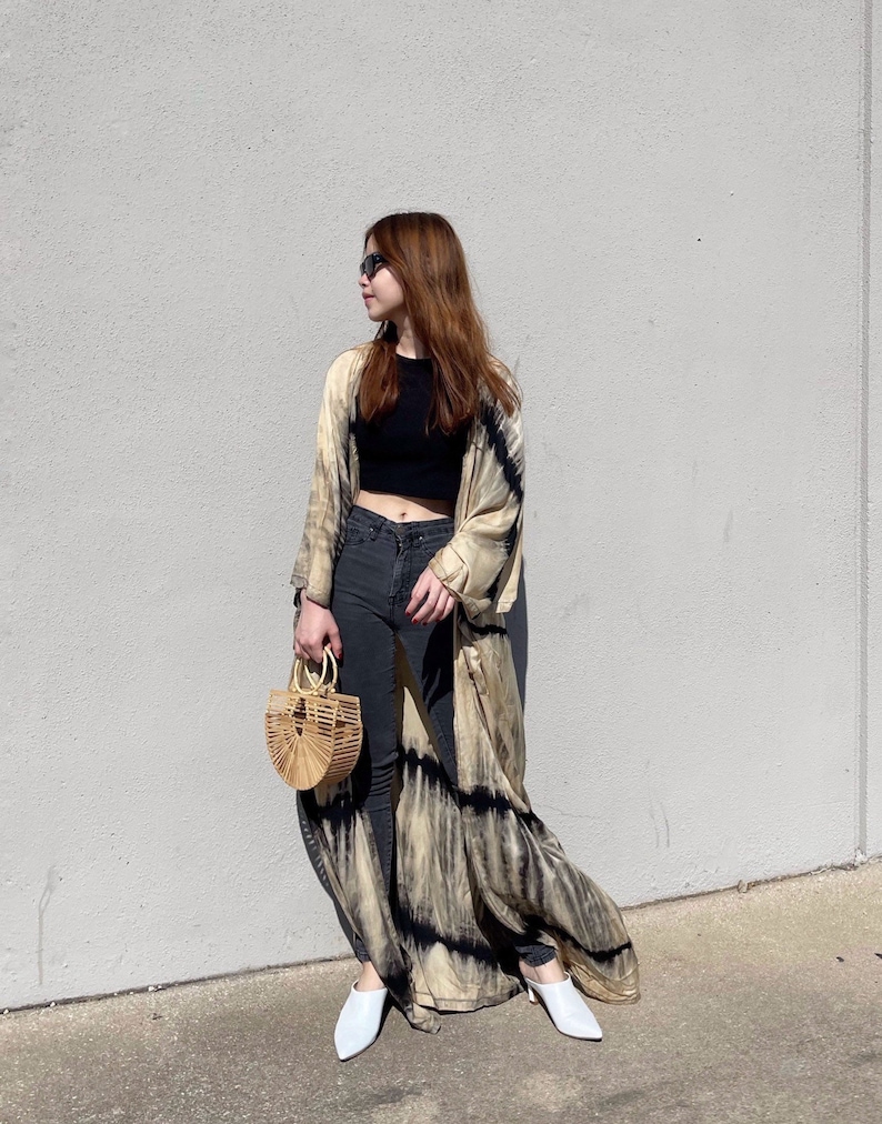 May include: A woman wearing a long, flowing tie-dye kimono with black accents over a black crop top and dark wash jeans. She is carrying a small, round, woven bag and wearing white shoes.