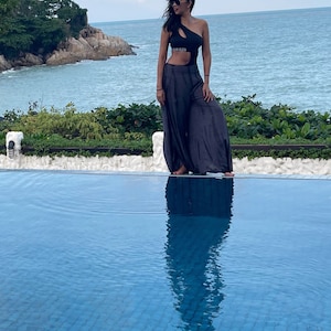 Puede incluir: Una mujer con un traje de ba&ntilde;o negro de una pieza con recortes y un logotipo de Calvin Klein se encuentra junto a una piscina. La piscina est&aacute; rodeada por un borde de azulejos blancos y exuberante vegetaci&oacute;n. La mujer lleva gafas de sol y tiene el pelo largo y oscuro.