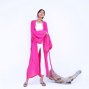 May include: A woman wearing a pink and orange tie-dye kimono with long sleeves and a white tank top. She is standing on a white background with a piece of driftwood in front of her.
