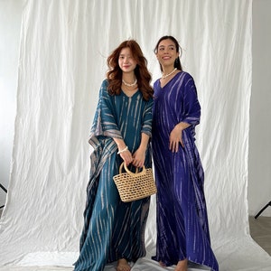 May include: Two women wearing long, flowing dresses with a V-neck design. One dress is teal with white stripes, and the other is a deep blue with white stripes. One woman holds a small, woven basket. The dresses appear to be made of a lightweight fabric.