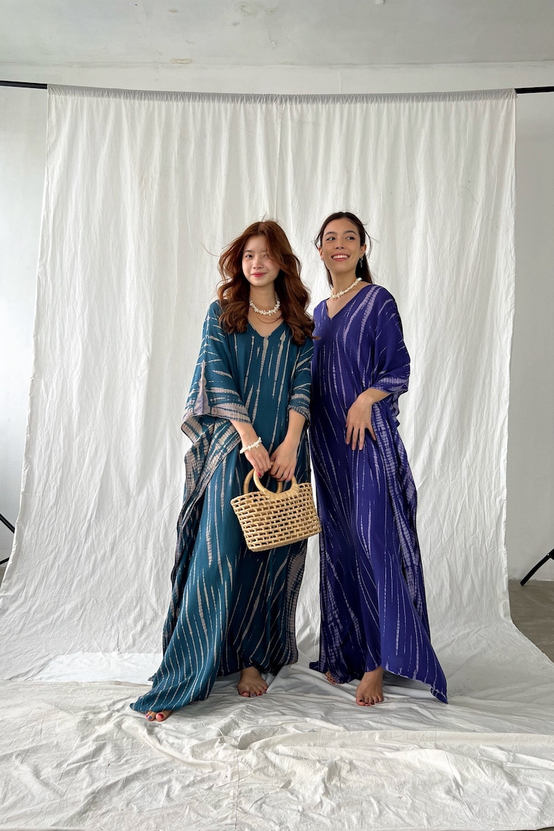 May include: Two women wearing long, flowing dresses with a V-neck design. One dress is teal with white stripes, and the other is a deep blue with white stripes. One woman holds a small, woven basket. The dresses appear to be made of a lightweight fabric.