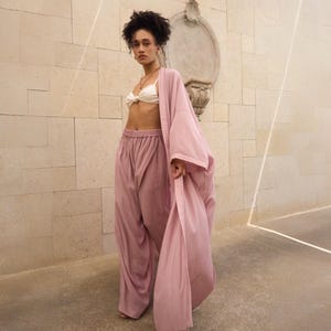 Op de afbeelding: Een oudroze open kamerjas met lange mouwen en een bijpassende wijde broek met een elastische tailleband. De outfit is gecombineerd met een wit geknoopt crop-topje, wat een set vormt van comfortabele, vloeiende loungewear-kledingstukken.