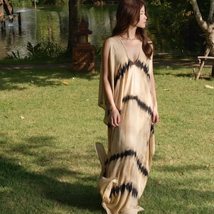 May include: A woman wearing a long, flowing, beige dress with black tie-dye patterns. The dress has a V-neckline and a loose, comfortable fit. She is standing in a grassy area with a pond in the background.