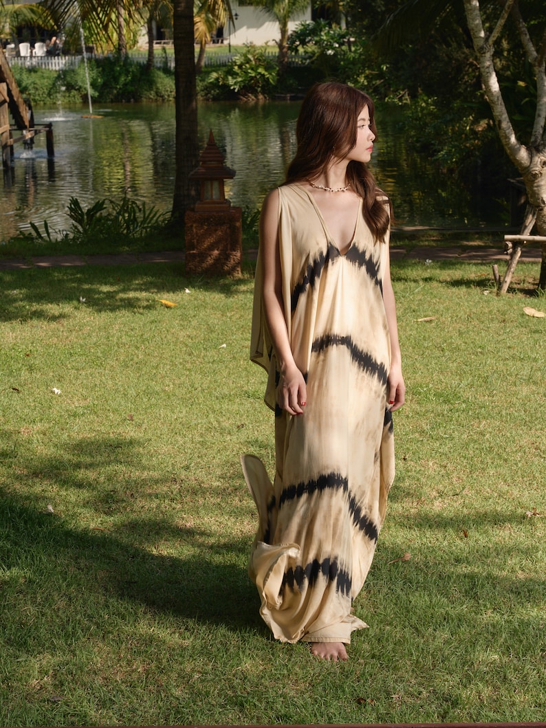 May include: A woman wearing a long, flowing, beige dress with black tie-dye patterns. The dress has a V-neckline and a loose, comfortable fit. She is standing in a grassy area with a pond in the background.