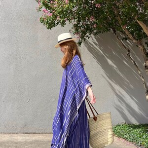 May include: A woman wearing a blue and white striped caftan, a straw hat, and carrying a straw tote bag with brown leather handles. The caftan is floor-length. The background includes a gray wall and a tree with pink flowers.