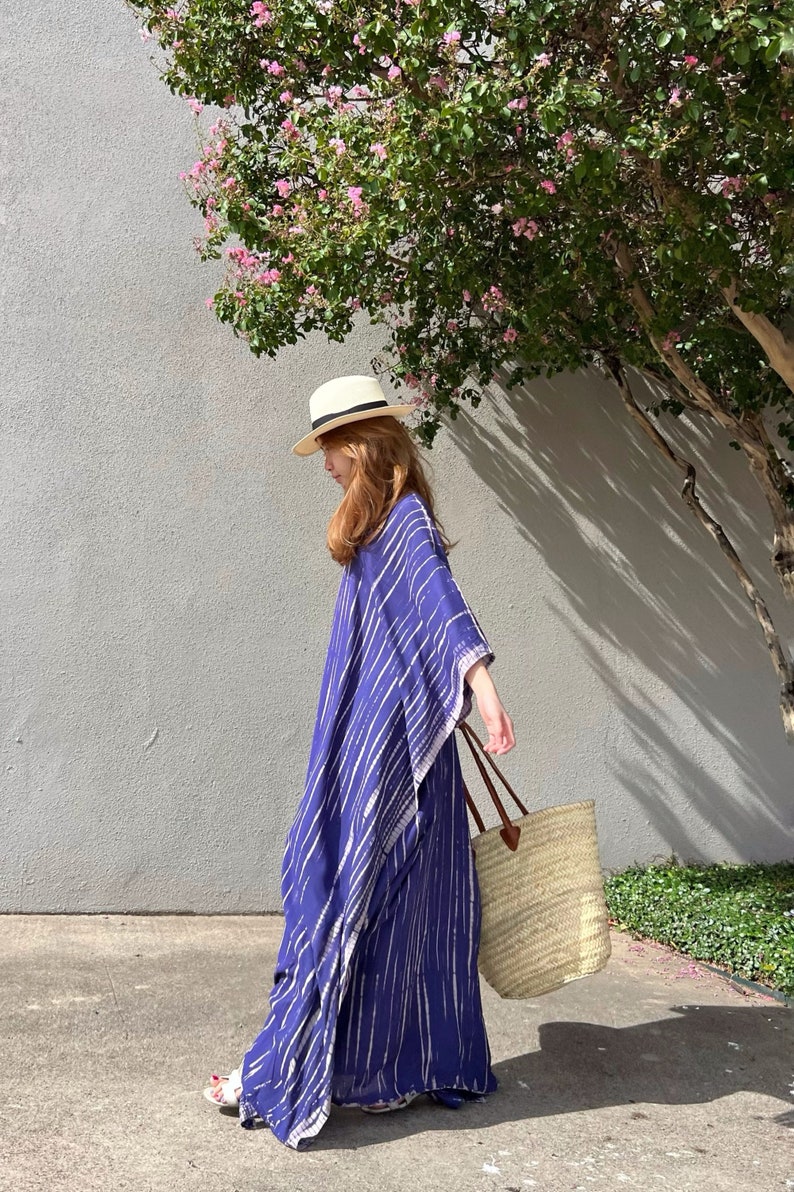 May include: A woman wearing a blue and white striped caftan, a straw hat, and carrying a straw tote bag with brown leather handles. The caftan is floor-length. The background includes a gray wall and a tree with pink flowers.