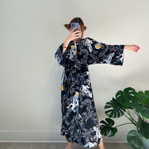 May include: A black kimono-style robe with a crane and foliage pattern. The robe features long sleeves and a floor-length design. The cranes are white, blue, and yellow. The robe is worn with white sandals.