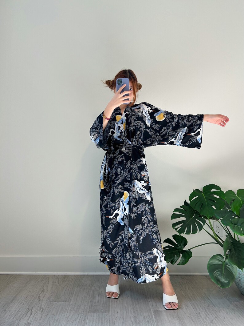 May include: A black kimono-style robe with a crane and foliage pattern. The robe features long sleeves and a floor-length design. The cranes are white, blue, and yellow. The robe is worn with white sandals.