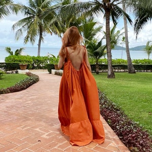 model in burn orange open back maxi dress with a low back and tie straps. The maxi dress has a tiered skirt and is being carried with a small woven handbag. The person is walking on a brick pathway with a tropical background.