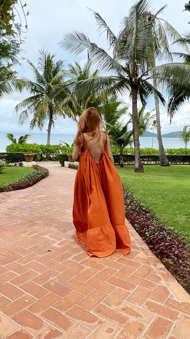 model in burn orange open back maxi dress with a low back and tie straps. The maxi dress has a tiered skirt and is being carried with a small woven handbag. The person is walking on a brick pathway with a tropical background.