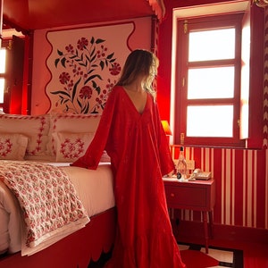 Op de afbeelding: Een vrouw in een rode jurk staat in een rode kamer met een bed en een raam. De kamer heeft wit-rode gestreepte behang en een wit-rood gepatroneerd tapijt. Het raam heeft een glas-in-loodontwerp.