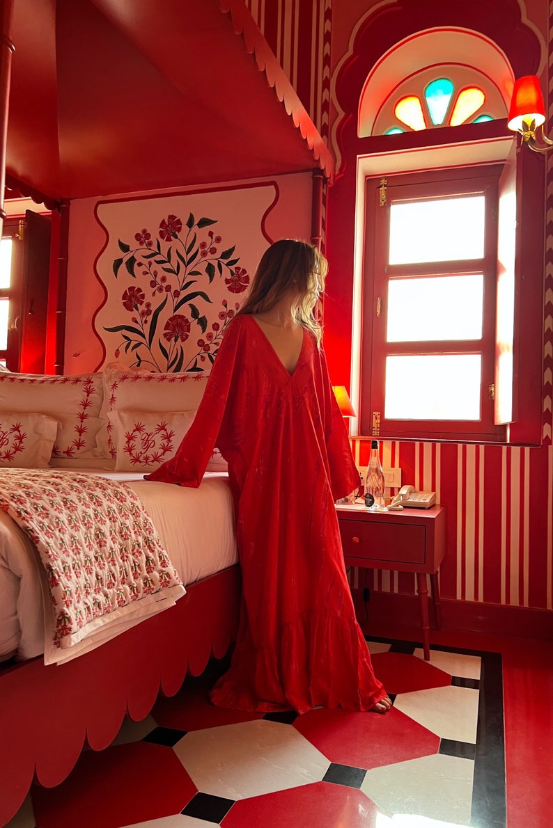 Op de afbeelding: Een vrouw in een rode jurk staat in een rode kamer met een bed en een raam. De kamer heeft wit-rode gestreepte behang en een wit-rood gepatroneerd tapijt. Het raam heeft een glas-in-loodontwerp.