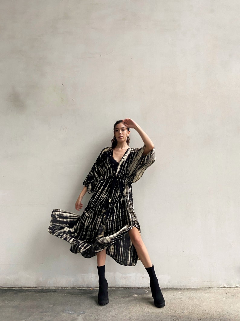 May include: A woman wearing a black and white tie-dye maxi dress with a long sleeve and a wrap style. She is wearing black ankle boots and has her hair pulled back.