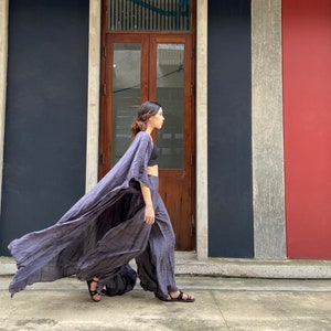 Puede incluir: Una mujer con un vestido largo, fluido, gris y drapeado con un top y pantalones negros. Est&aacute; caminando frente a un edificio con una puerta de madera y una pared roja.
