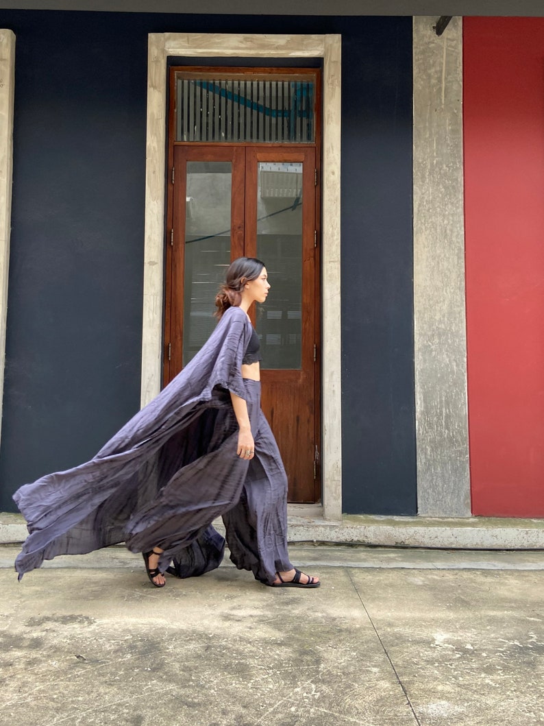 Puede incluir: Una mujer con un vestido largo, fluido, gris y drapeado con un top y pantalones negros. Est&aacute; caminando frente a un edificio con una puerta de madera y una pared roja.