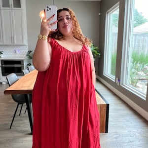 May include: A woman wearing a red maxi dress with a scoop neck and thin straps. The dress has a gathered bodice and a flowing skirt.