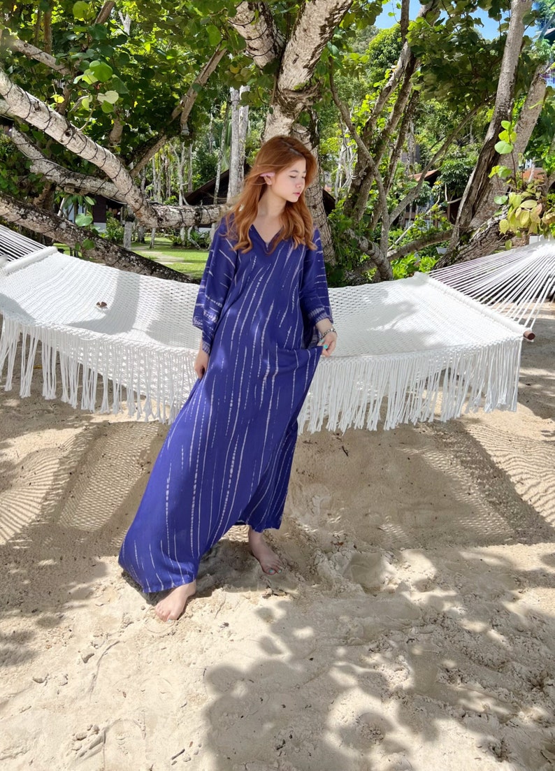 May include: A long, flowing blue dress with white vertical stripes. The dress has a V-neck and wide sleeves. The person is standing on a sandy beach near a white hammock, under the shade of a tree.