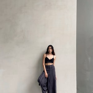 Puede incluir: Una mujer con un sujetador de encaje negro y pantalones anchos de color gris oscuro. Est&aacute; de pie frente a una pared blanca con una pared de acento gris a la derecha.