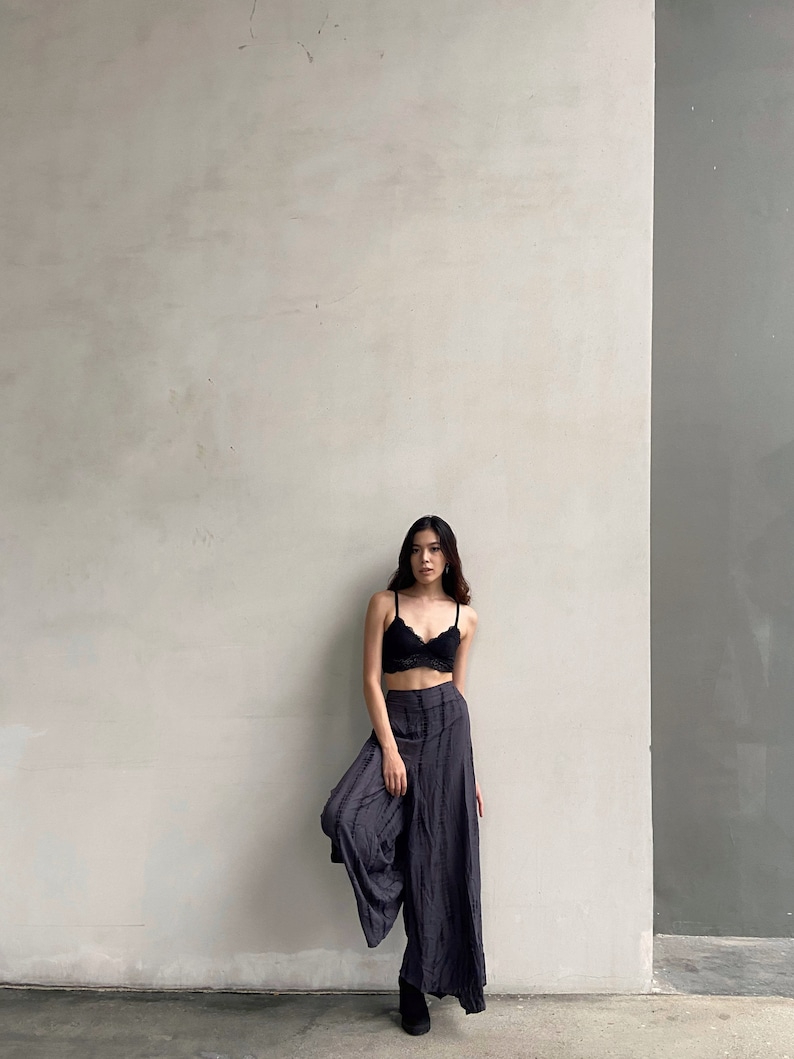 Puede incluir: Una mujer con un sujetador de encaje negro y pantalones anchos de color gris oscuro. Est&aacute; de pie frente a una pared blanca con una pared de acento gris a la derecha.