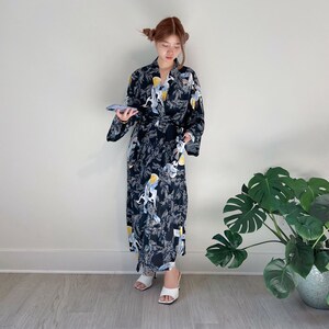 May include: A black kimono-style robe with a repeating pattern of white and yellow birds and foliage. The robe is belted at the waist and worn with white sandals. The background is a plain white wall.