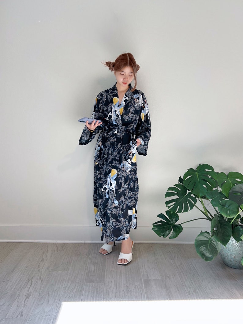 May include: A black kimono-style robe with a repeating pattern of white and yellow birds and foliage. The robe is belted at the waist and worn with white sandals. The background is a plain white wall.
