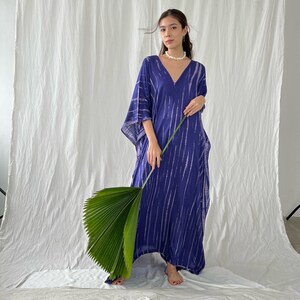 May include: A long, flowing blue dress with a V-neck and white vertical stripe pattern. The dress has wide sleeves and is worn by a person holding a large green leaf. The background is a white sheet.