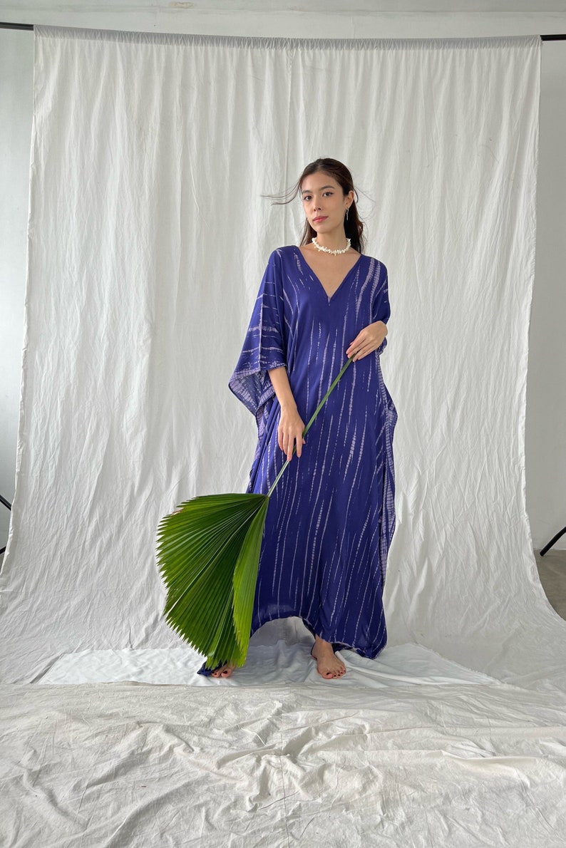 May include: A long, flowing blue dress with a V-neck and white vertical stripe pattern. The dress has wide sleeves and is worn by a person holding a large green leaf. The background is a white sheet.