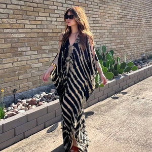 May include: A long, flowing dress with a black and beige striped pattern. The dress has a V-neck and open sleeves. The person is wearing sunglasses and brown sandals. The background includes a brick wall and a sunny outdoor setting.