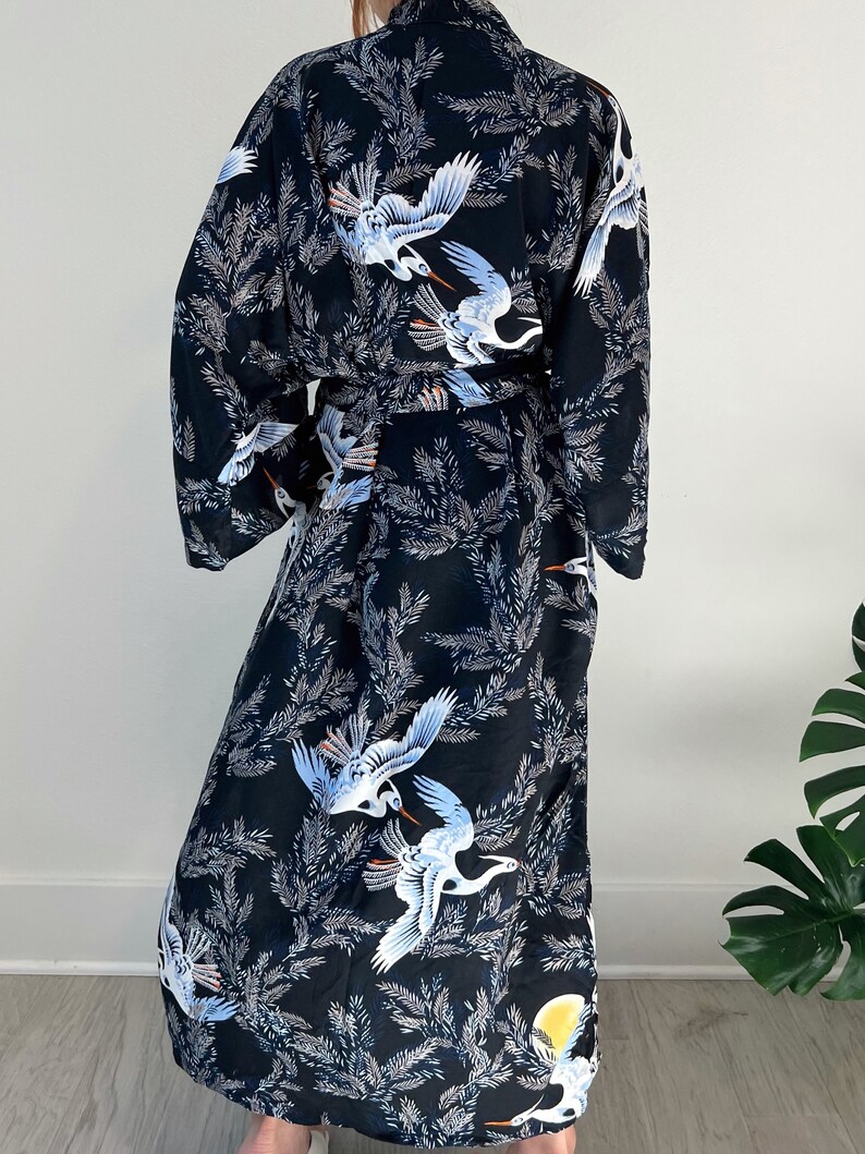 May include: Black kimono robe featuring a repeating pattern of white and blue cranes and foliage. The robe has long sleeves and a matching belt. The background is a light color.