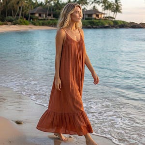 A rust-colored oprn back maxi dress with spaghetti straps and a ruffled hem. The dress is worn on a beach with the ocean in the background. Palm trees and buildings are visible in the distance. The dress is made of a lightweight fabric.