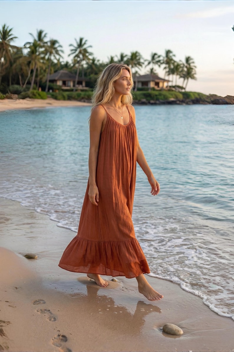 A rust-colored oprn back maxi dress with spaghetti straps and a ruffled hem. The dress is worn on a beach with the ocean in the background. Palm trees and buildings are visible in the distance. The dress is made of a lightweight fabric.