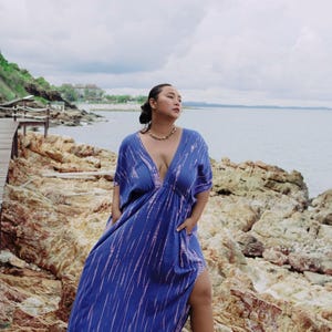 Op de afbeelding: Een vrouw draagt een blauwe en roze tie-dye maxi-jurk met een diepe V-hals en een zijsplit. Ze staat op een rotsachtig strand met de oceaan op de achtergrond.