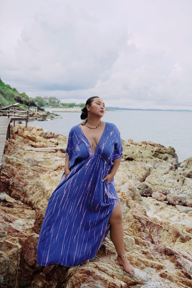Op de afbeelding: Een vrouw draagt een blauwe en roze tie-dye maxi-jurk met een diepe V-hals en een zijsplit. Ze staat op een rotsachtig strand met de oceaan op de achtergrond.