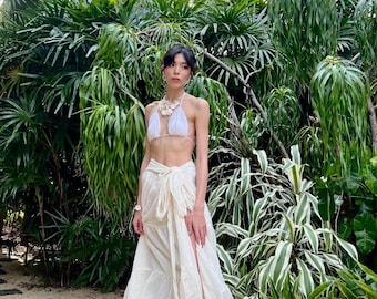 May include: A woman wearing a white bikini top and a white wrap skirt. She is standing in a tropical garden with lush green foliage.