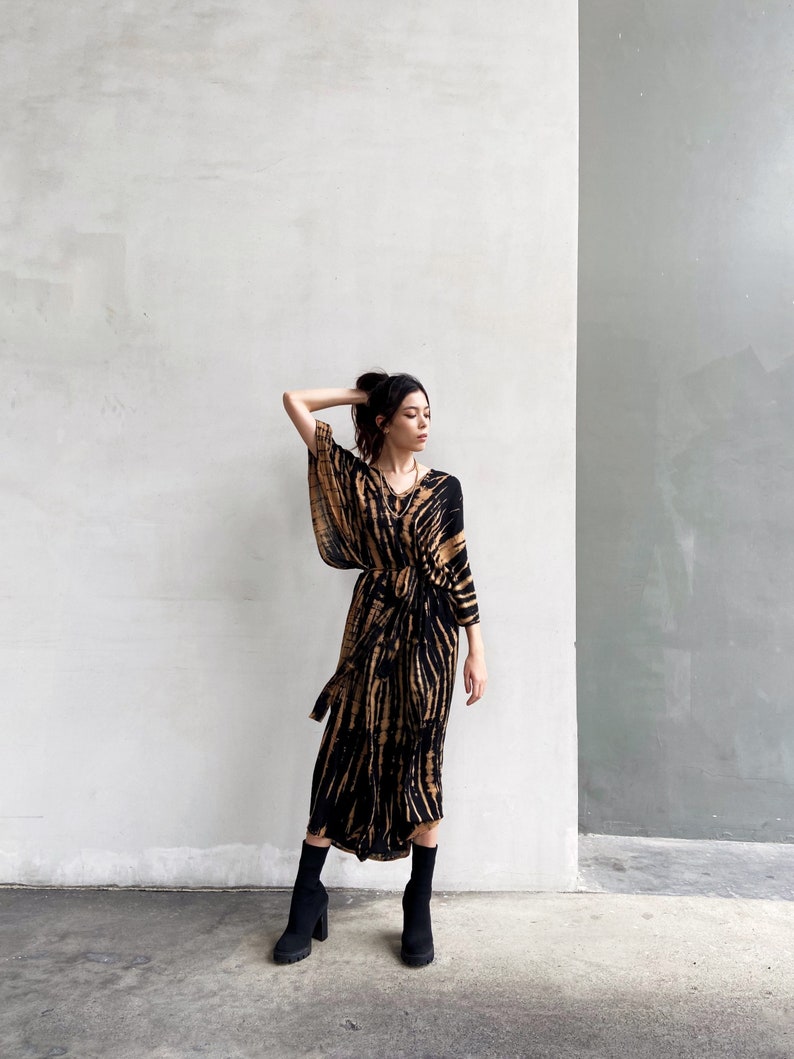 May include: A woman wearing a long, black and brown tie-dye dress with a long sleeve and a tie at the waist. She is wearing black ankle boots.