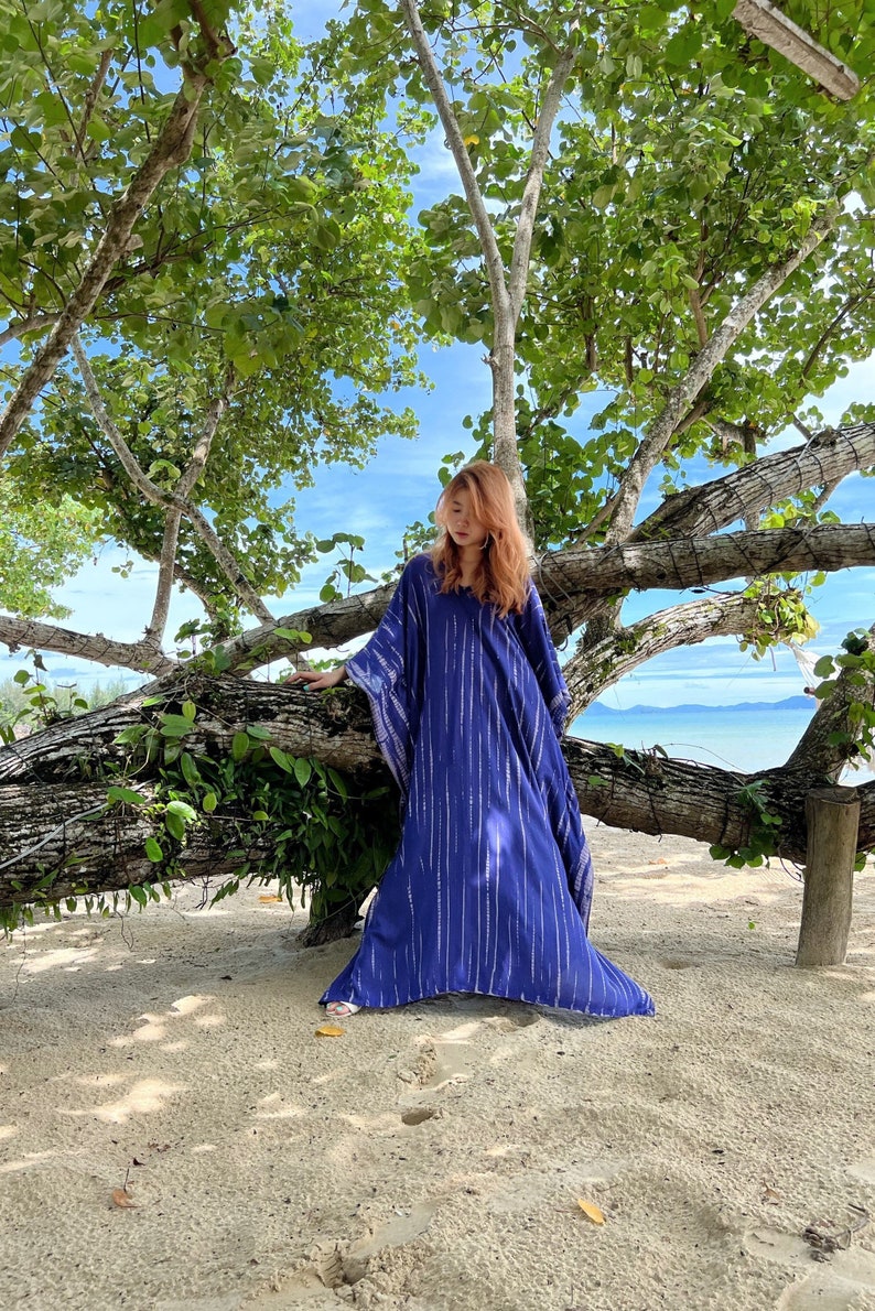 May include: A long, flowing, indigo blue caftan dress with vertical silver stripes. The dress has a V-neck and wide sleeves. The image is taken outdoors, with a beach and trees in the background.