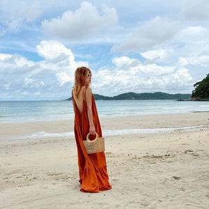 model in burn orange orn back maxi dress with a low back and tie straps. The maxi dress has a tiered skirt and is being carried with a small woven handbag. The person is walking on the beach.