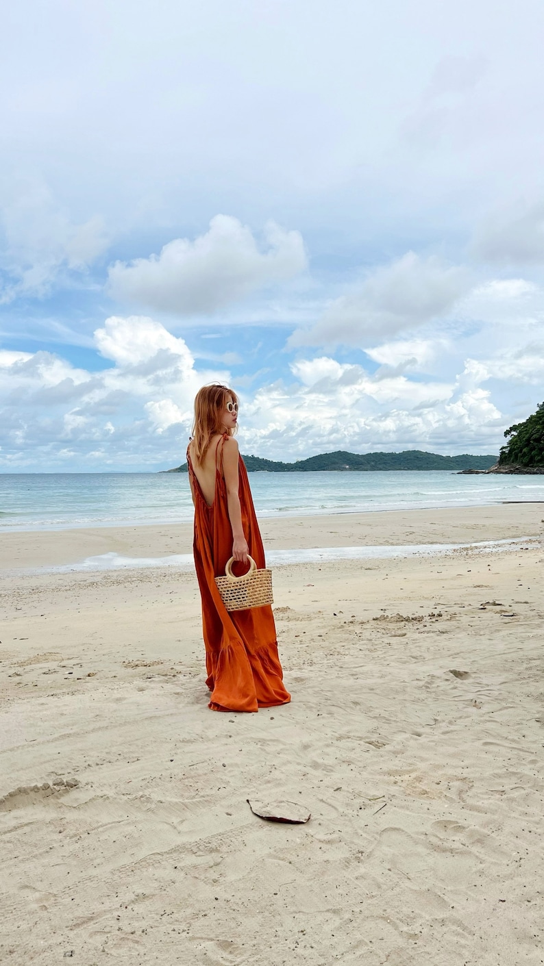 model in burn orange orn back maxi dress with a low back and tie straps. The maxi dress has a tiered skirt and is being carried with a small woven handbag. The person is walking on the beach.