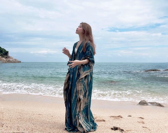 May include: A woman wearing a long, flowing, teal and brown patterned dress stands on a sandy beach with the ocean in the background.