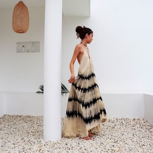 May include: A woman wearing a long, white maxi dress with black tie-dye patterns. The dress has a halter neckline and a flowing silhouette. She is standing on a gravel surface in front of a white wall.