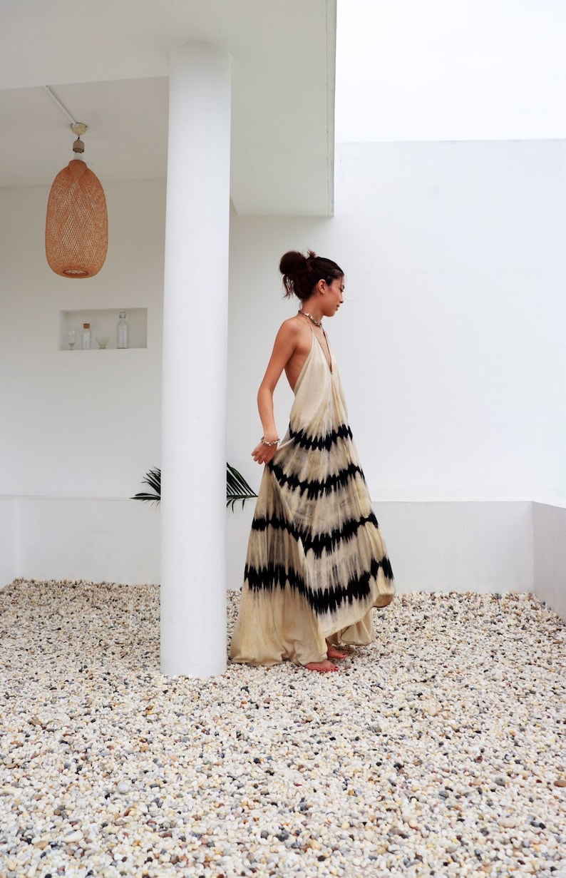 May include: A woman wearing a long, white maxi dress with black tie-dye patterns. The dress has a halter neckline and a flowing silhouette. She is standing on a gravel surface in front of a white wall.
